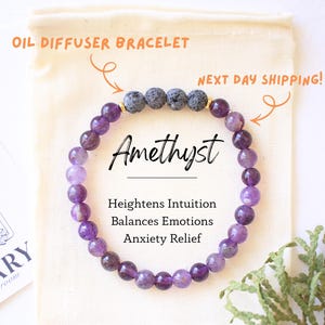 May include: Amethyst oil diffuser bracelet with purple beads and lava stones. The bracelet is on a cream-colored pouch with the word "Amethyst" and the benefits of the stone. A card with a floral design is on the left.