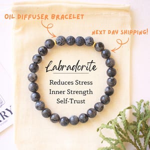 Puede incluir: Una pulsera difusora de aceites de labradorita con cuentas grises. La pulsera se muestra en una bolsa beige con el texto "Labradorita Reduce el estrés Fuerza interior Confianza en sí mismo". También hay una tarjeta con ilustraciones florales.