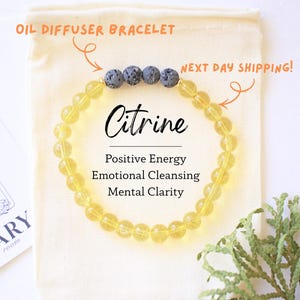 May include: A Citrine oil diffuser bracelet with yellow beads and dark gray lava stone accents. The bracelet is displayed on a cream-colored pouch with text that reads "Citrine: Positive Energy, Emotional Cleansing, Mental Clarity."