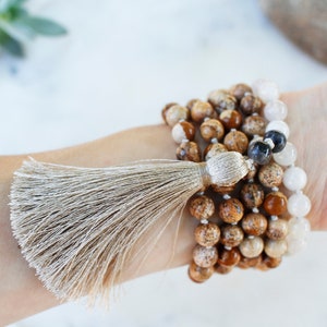 Pode incluir: Um bracelete de v&aacute;rias voltas feito de contas marrons, brancas e pretas com um tassel bege. O bracelete &eacute; usado no pulso.