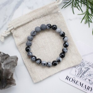 May include: A gray and black gemstone bracelet with a natural stone look. The bracelet is on a white fabric pouch with the text "the ROSEMARY" printed on a card.