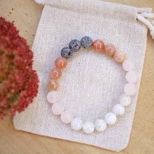 May include: A bracelet made with natural stones in shades of white, pink, and orange. The bracelet features lava rock beads and is displayed on a white fabric pouch.