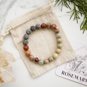 May include: A multi-colored gemstone bracelet with lava rock beads. The bracelet is in a small, white drawstring bag. The bag is on a white marble surface. A small card with the text "the ROSEMARY room" is in the background.