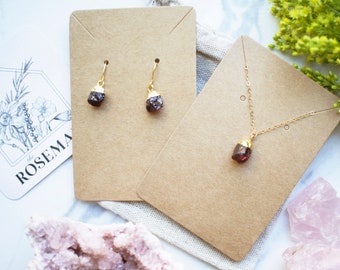 May include: A set of gold-plated earrings and a necklace featuring raw, dark red garnet crystals. The jewelry is displayed on brown card stock with a white background.