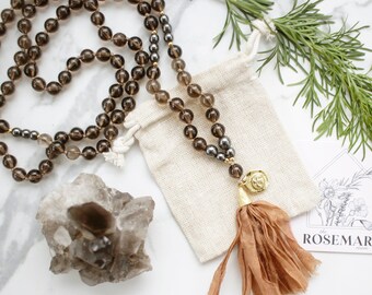 May include: A brown and gold beaded mala necklace with a tassel and a small pouch. The necklace is made of smoky quartz and hematite beads. The pouch is made of natural fabric. The image also includes a small crystal cluster and a card with the text "the ROSEMARY project".