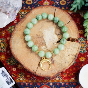 May include: A green jade stone bracelet with a gold crescent moon charm. The bracelet is on a wooden surface with a red and gold patterned background.