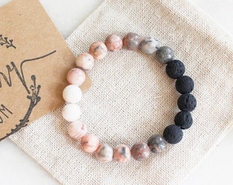 May include: A pink zebra jasper and black lava rock bracelet with a white bead. The bracelet is in a small, white drawstring bag. The bag has a brown tag that says "The Rosemary Room" and "Best Seller".