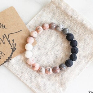 May include: A pink zebra jasper and black lava rock bracelet with a white bead. The bracelet is in a small, white drawstring bag. The bag has a brown tag that says "The Rosemary Room" and "Best Seller".