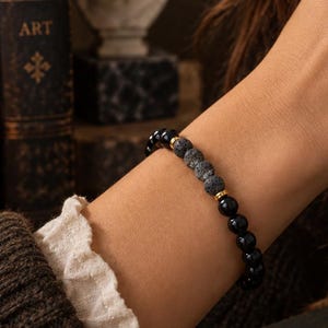 Puede incluir: Una pulsera de cuentas que presenta cuentas negras redondas y lisas alternadas con cuentas de piedra de lava gris texturizadas y porosas, acentuadas por pequeños espaciadores de metal dorado.