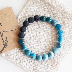 Puede incluir: Un brazalete de piedras azules y negras con cuentas de lava. El brazalete está en una bolsa de tela blanca con el texto "The Rosemary Room" y "Best Seller" en una tarjeta marrón.