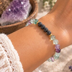 May include: A beaded bracelet featuring smooth, round, multi-colored fluorite stones in shades of green and purple, interspersed with three textured black lava beads and small gold-toned metal spacers, worn on a wrist with a white textured sleeve.