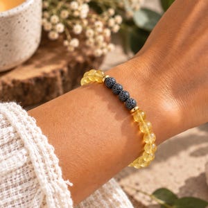 Puede incluir: Una pulsera de cuentas con esferas de piedras preciosas amarillas translúcidas y una sección central de cuatro cuentas de piedra de lava gris oscuro porosa, acentuada por pequeños espaciadores de metal dorado, sobre una muñeca con manga blanca.