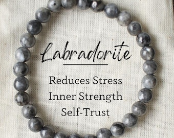 May include: A gray Labradorite gemstone bracelet on a white fabric pouch. The pouch has the text "Labradorite", "Reduces Stress", "Inner Strength", and "Self-Trust" printed on it.