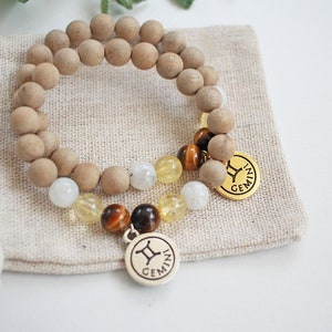 May include: Two beaded bracelets with wooden beads, gemstones, and gold-colored charms. One bracelet features a Gemini zodiac charm. The bracelets rest on a small beige fabric pouch, ideal for gifting or storage.