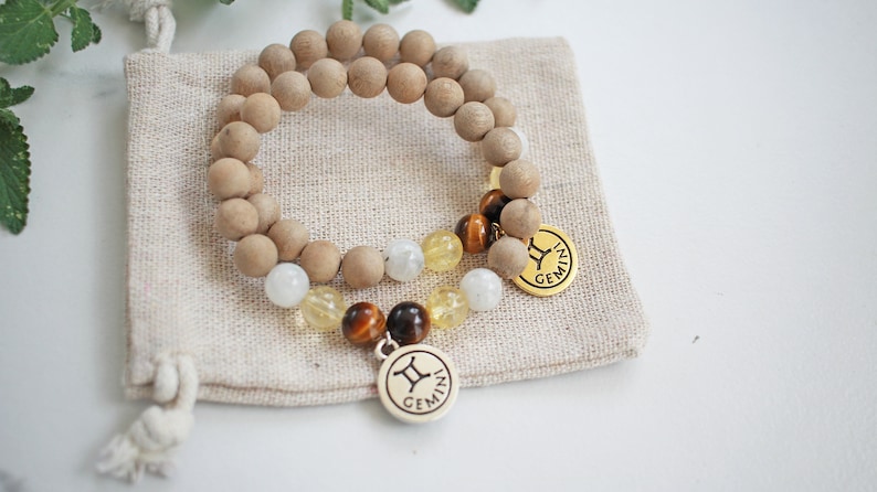 May include: Two beaded bracelets with wooden beads, gemstones, and gold-colored charms. One bracelet features a Gemini zodiac charm. The bracelets rest on a small beige fabric pouch, ideal for gifting or storage.