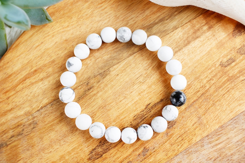 May include: White and black beaded bracelet with a single black bead. The bracelet is made of round, smooth beads.