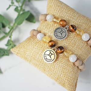 May include: Two beaded bracelets with Gemini charms rest on a small burlap pillow. The bracelets feature wooden beads, white and yellow gemstones, and round charms with the Gemini symbol and the word "GEMINI".