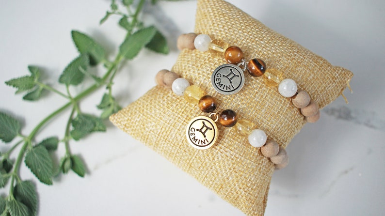 May include: Two beaded bracelets with Gemini charms rest on a small burlap pillow. The bracelets feature wooden beads, white and yellow gemstones, and round charms with the Gemini symbol and the word "GEMINI".