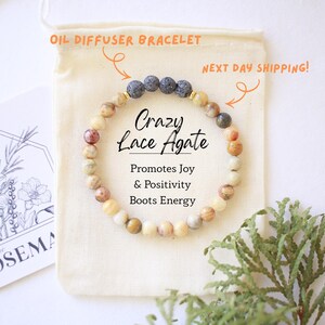 Pode incluir: Uma pulseira difusora de óleos de ágata Crazy Lace. A pulseira é apresentada em uma bolsa creme, com as palavras "Crazy Lace Agate" e "Promotes Joy & Positivity Boots Energy". Um cartão com ilustrações florais também é visível.