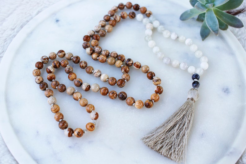 Pode incluir: Um colar mala de pedras preciosas marrons e brancas com um tassel. O colar &eacute; feito de contas redondas e tem uma conta cinza escura no final.