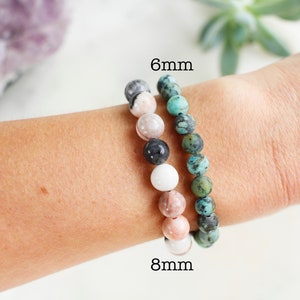 May include: Two beaded bracelets on a wrist. The bracelet on the left is made of 8mm beads in shades of pink, white, and black. The bracelet on the right is made of 6mm turquoise beads.