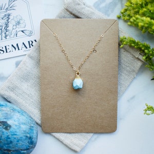 Puede incluir: Un delicado collar de cadena dorada con un colgante geom&eacute;trico azul claro. El collar se muestra en una tarjeta marr&oacute;n con fondo blanco.