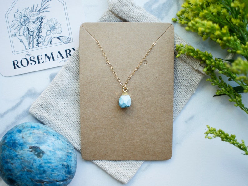Puede incluir: Un delicado collar de cadena dorada con un colgante geom&eacute;trico azul claro. El collar se muestra en una tarjeta marr&oacute;n con fondo blanco.