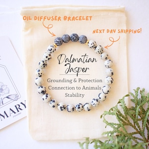 May include: Dalmatian Jasper oil diffuser bracelet. The bracelet features round, speckled beads in white and black, with a few gray beads. The text on the image says "Grounding & Protection, Connection to Animals, Stability."