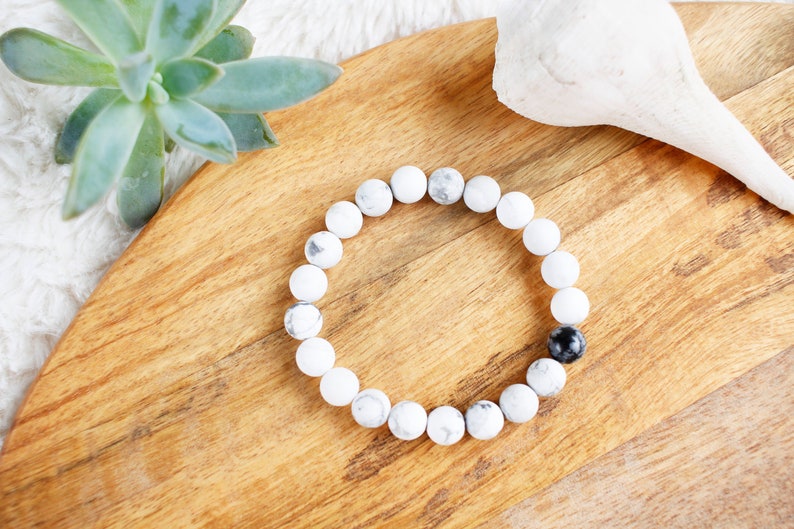 May include: A white and black beaded bracelet with a single black bead. The bracelet is on a wooden surface.
