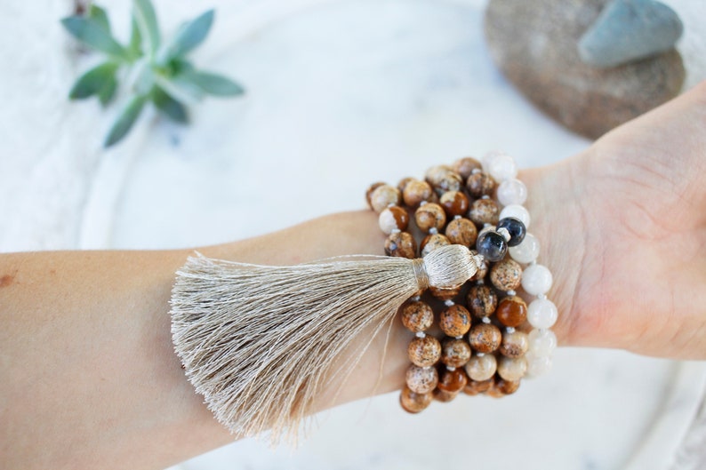 Pode incluir: Um bracelete de v&aacute;rias voltas feito de contas marrons, brancas e pretas com um tassel bege. O bracelete &eacute; usado no pulso.