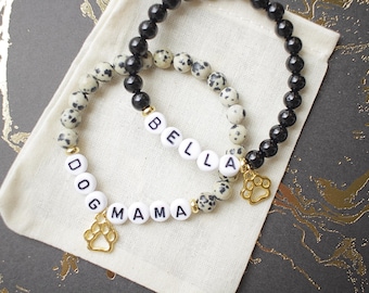 Könnte beinhalten: Zwei Armbänder mit schwarzen und weißen gesprenkelten Perlen und goldenen Pfotenanhängern. Ein Armband buchstabiert "DOG MAMA" und das andere "BELLA".