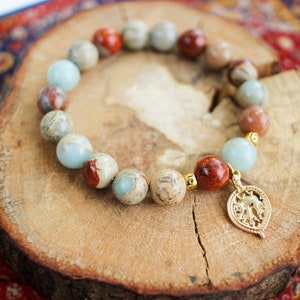 May include: A bracelet made of brown, beige, and blue stones with a gold elephant charm. The bracelet is on a wooden surface.
