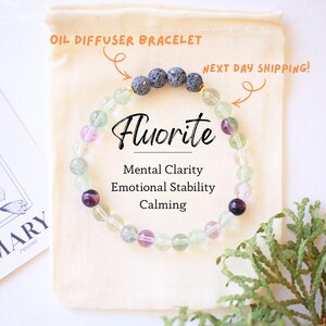 May include: Fluorite oil diffuser bracelet with green, purple, and gray beads. The bracelet is on a beige pouch with the word "Fluorite" in black script. Text on the pouch says "Mental Clarity, Emotional Stability, Calming."