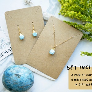 Puede incluir: Un conjunto de joyas de piedra preciosa azul claro sobre cartulina marr&oacute;n. El conjunto incluye un par de pendientes y un collar a juego. El texto "SET INCLUDES: A PAIR OF EARRINGS & A MATCHING NECKLACE IN GIFT WRAPPING" est&aacute; impreso sobre un fondo amarillo.