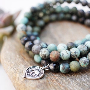 May include: A multi-strand turquoise and black beaded bracelet with a silver elephant charm. The bracelet is wrapped around a piece of wood.