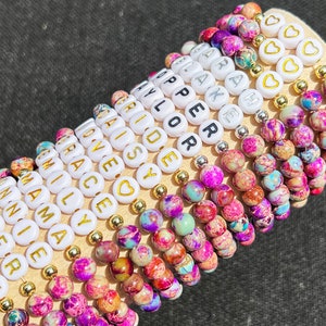May include: A stack of colourful bracelets with white beads that spell out names. The bracelets are made with small, colourful, speckled beads.