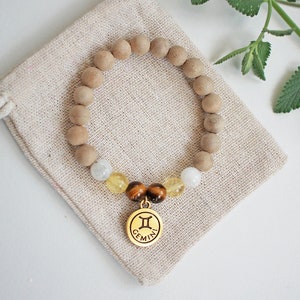 May include: A beaded bracelet with wooden beads, gemstones, and a gold-colored Gemini charm. The bracelet rests on a beige drawstring pouch. Green leaves are in the background. The bracelet is a stylish accessory.