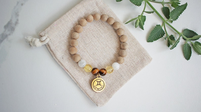 May include: A beaded bracelet with wooden beads, gemstones, and a gold-colored Gemini charm. The bracelet rests on a beige drawstring pouch. Green leaves are in the background. The bracelet is a stylish accessory.