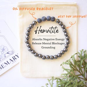 May include: Hematite oil diffuser bracelet with dark gray beads. The bracelet is displayed on a small beige pouch, with text indicating it absorbs negative energy and releases mental blockages. A card with floral illustrations and text is also visible.