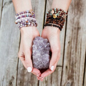 Pode incluir: Um par de m&atilde;os segurando um grande cristal de ametista. O cristal &eacute; de cor roxo escuro e tem muitas facetas. A pessoa est&aacute; usando v&aacute;rias pulseiras em ambos os pulsos.