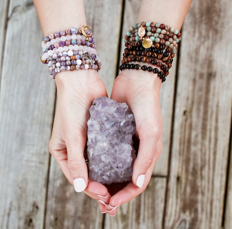 Pode incluir: Um par de m&atilde;os segurando um grande cristal de ametista. O cristal &eacute; de cor roxo escuro e tem muitas facetas. A pessoa est&aacute; usando v&aacute;rias pulseiras em ambos os pulsos.