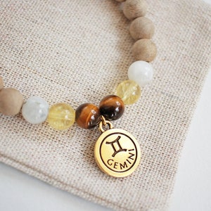 May include: A beaded bracelet featuring light brown wooden beads, white and yellow gemstone beads, and a gold-colored charm. The charm displays the Gemini zodiac symbol and the word "GEMINI". The bracelet rests on a beige linen surface.