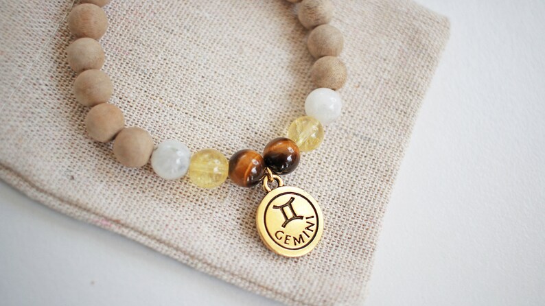 May include: A beaded bracelet featuring light brown wooden beads, white and yellow gemstone beads, and a gold-colored charm. The charm displays the Gemini zodiac symbol and the word "GEMINI". The bracelet rests on a beige linen surface.