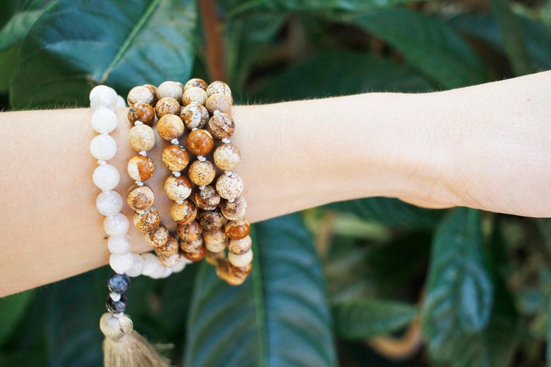 Pode incluir: Um conjunto de quatro pulseiras de contas, duas s&atilde;o brancas e duas s&atilde;o marrons com manchas brancas e marrons. As pulseiras est&atilde;o empilhadas no pulso de uma pessoa.