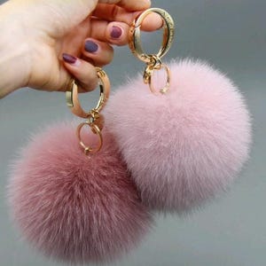 May include: Two fluffy pink fur pom-poms with gold-colored keychains. The pom-poms are different shades of pink, one a deeper rose color and the other a lighter pastel pink. The keychains have a circular clasp and a small ring.