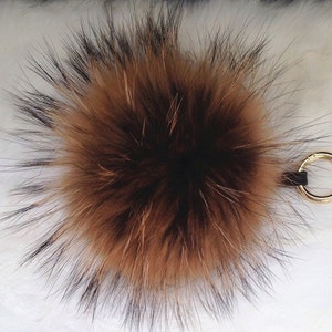May include: A fluffy, round keychain accessory made of brown and black fur. The fur is dense and has a natural, textured appearance. A gold-colored metal ring and a small leather strap are attached for easy hanging.