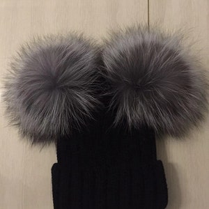 May include: Black knitted beanie hat featuring two large, fluffy, grey fur pom-poms. The hat is made of a ribbed knit material. The pom-poms are a light grey colour, adding a touch of luxury to the winter hat.