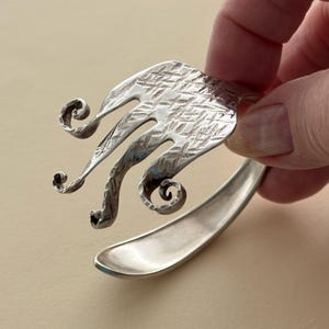 May include: A silver-toned metal cuff bracelet crafted from a repurposed fork. The tines of the fork are curled outward into decorative spirals, and the upper portion of the fork features a textured, cross-hatched pattern on the metallic surface.