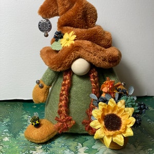 May include: A plush gnome with a brown hat and green outfit holding a bouquet of sunflowers and other fall flowers. The gnome has a wooden nose and a braided beard. There is a small bee on the gnome's foot.