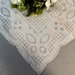 May include: A white lace handkerchief with a delicate floral pattern. The handkerchief is decorated with a repeating geometric design and has a scalloped edge.
